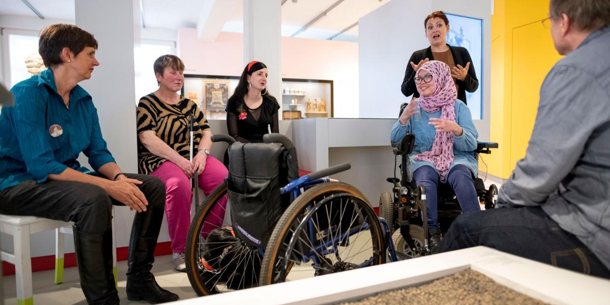 Sechs Menschen im Ausstellungsbereich. Sie sitzen einander zugewandt. Eine junge Frau mit Kopftuch sitzt im Rollstuhl. Sie spricht lächelt. Hinter ihr steht eine Gebärdensprachdolmetscherin und übersetzt. In der Bildmitte ein leerer Rollstuhl.