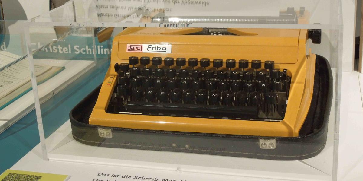 Eine sonnengelbe mechanische Schreibmaschine unter einer Acrylhaube auf dem Ausstellungstisch. 
