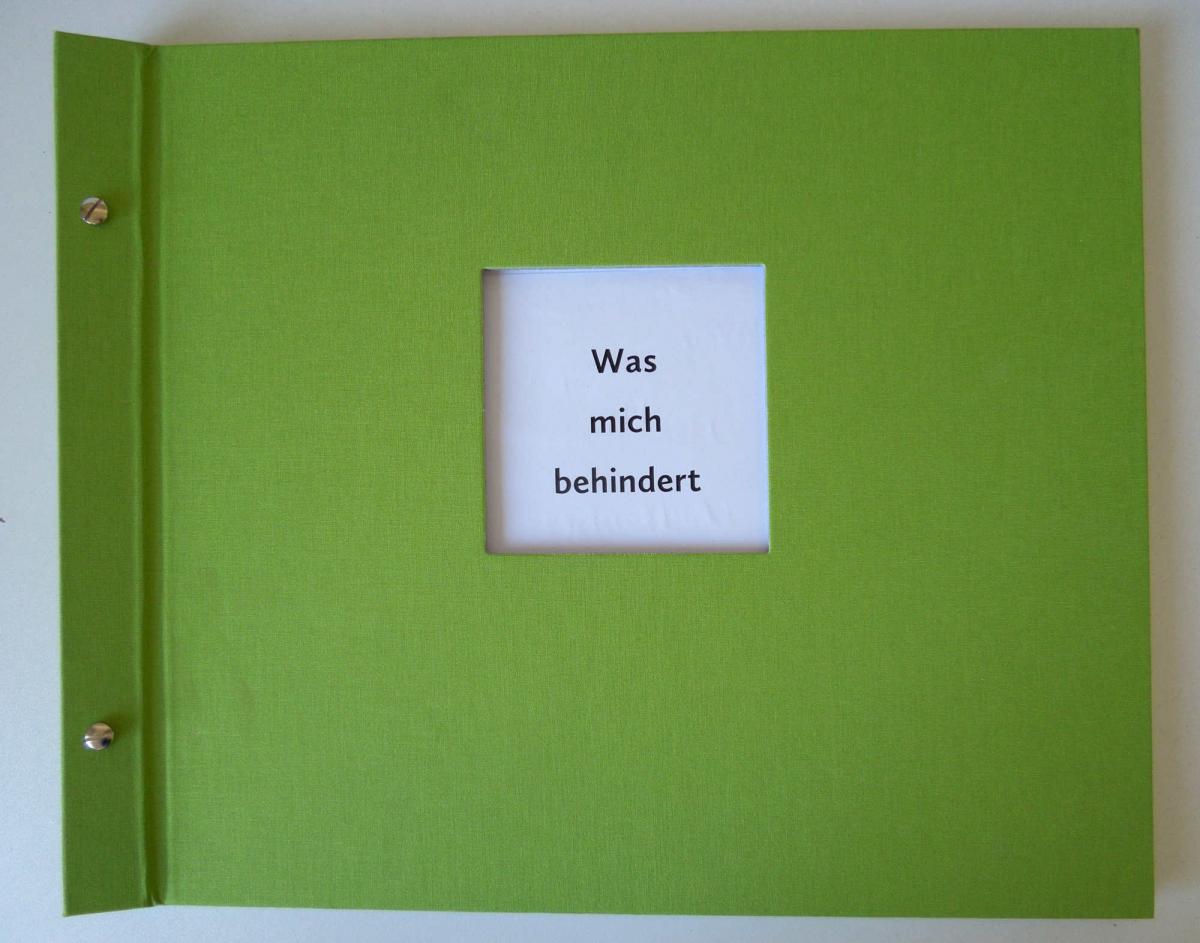 In grünen Leinen gebundenes Buch mit einer quadratischen Öffnung auf dem Deckel. Darin der Text: Was mich behindert