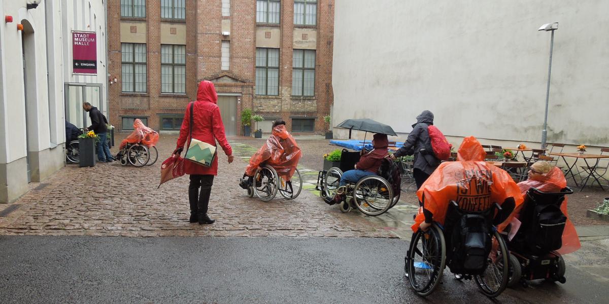 Im Hof des Stadtmuseums. Eine Gruppe Rollstuhlfahrer fährt hintereinander zum Eingang.Es regnet. Die meisten tragen Regencapes.