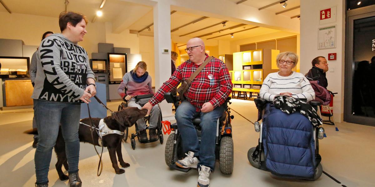 Menschen im der Ausstellung des Stadtmuseums. Eine Frau mit Blindenhund und ein Mann im Rollstuhl unterhalten sich lachend miteinander. Eine andere Frau im Rollstuhl sitzt daneben. Im Hintergrund weitere Menschen. 