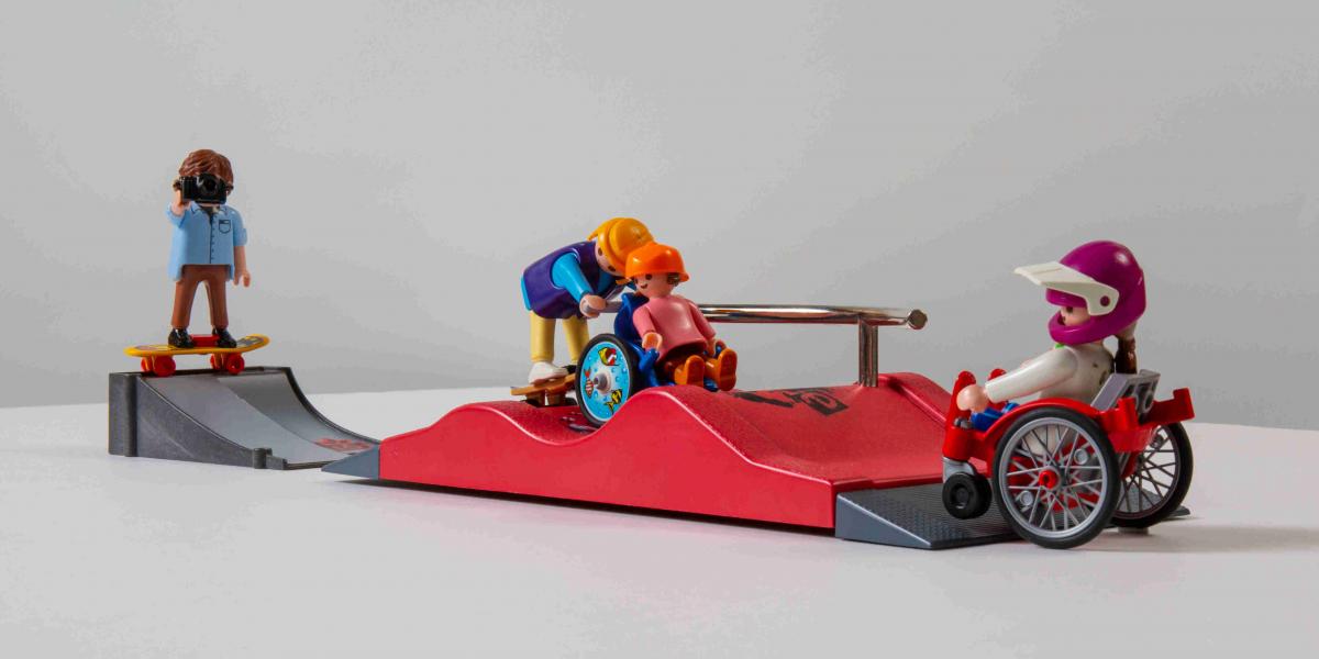 Vier Playmobilfiguren auf zwei Rampen. Ein Kind im Rollstuhl wird von einer Skaterin geschoben. Eine Frau im Rollstuhl am Fuß der Rampe. Ein Mann mit Skateboard fotografiert.