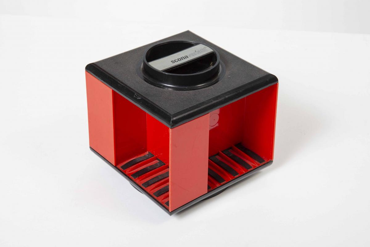 Rote Box mit schwarzem Deckel. Würfelform, drehbar gelagert. An vier Seiten sind Aussparungen für jeweils fünf Magnetbandkassetten.
