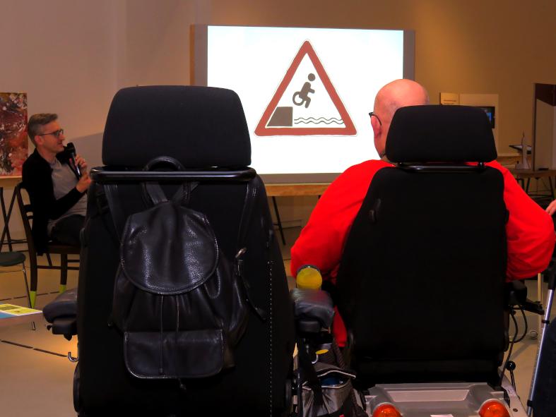 Zwei Menschen im Rollstuhl von hinten. Auf der Projektionsfläche vor Ihnen eine Zeichnung von einem ins Wasser stürzenden Rollstuhl - in Anlehnung an das Achtungsschild mit dem ins Wasser stürzenden Auto. Phil Hubbe links im Bild. Er spricht ins Mikrofon. 