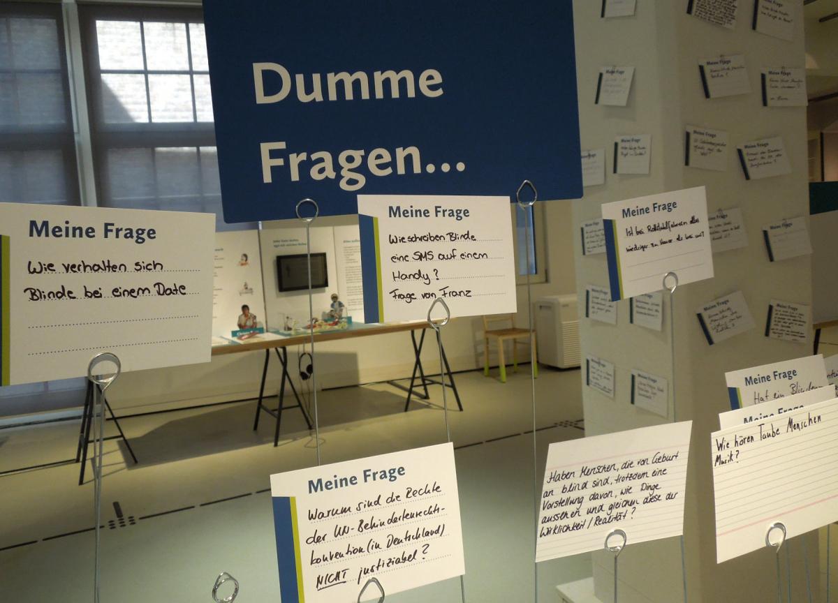 In der Ausstellung: Handgeschriebene Karten mit Fragen auf Ständern aus Draht, größeres Schild mit der Aufschrift "Dumme Fragen"