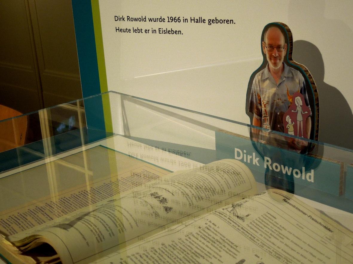 Austellungstisch mit aufgeschlagenem Ordner unter einer Acrylhaube, dahinter ein Foto von Dirk Rowold
