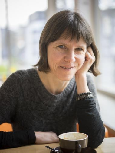 Die Autorin Dagmar Petrick an einem Tisch mit einer Tasse Kaffee. Sie stützt ihren Kopf auf den linken Arm und lächelt in die Kamera.